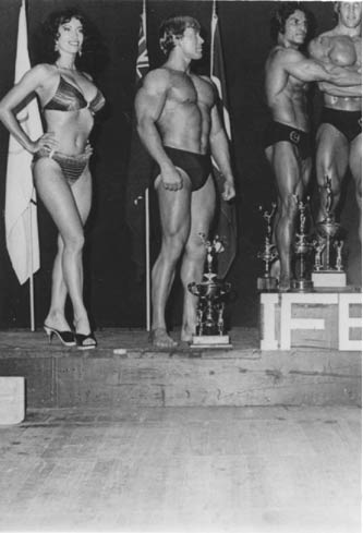Rock bodied muscle fem. Kelle Everts stands on the podium of Winner Circle at Bodybuilding competition. Muscle woman, hands on hips next to male hulks.