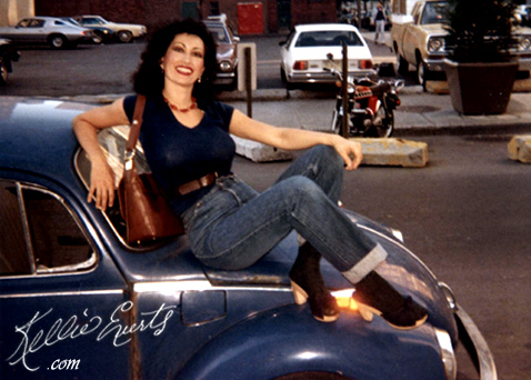 Kellie Everts. Sexy, curvy stripper, brown over shoulder purse, sittlng on hood of Volkswagon Beetle car in black boots and skin tight jeans. Montreal, Canada.