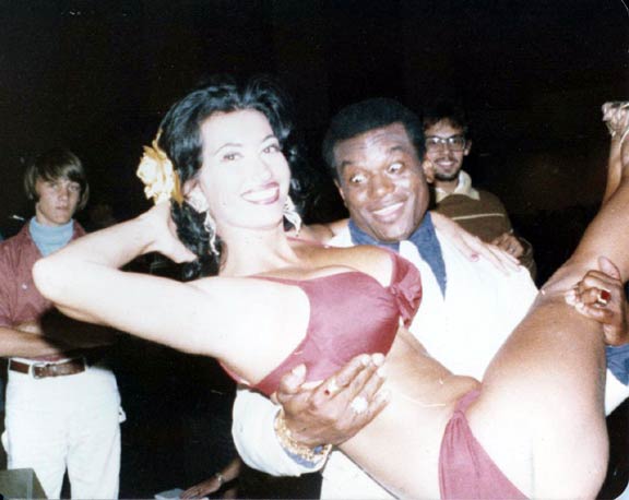 "Take a look at them apples!"  A bug-eyed & attentive Sergio Oliva carrying perfect body Kellie Everts at the 1975 Dan Lurie WBBG competition. Red, maroon bikini with rose in Kellie's black hair.
