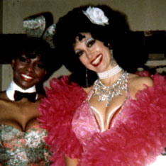 Sexy stripper with white rose in black permed hair standing next to her African American friend Joi, a fellow Playboy Bunny, flashes a perfect ten smile, dressed in beads, silver jewels, pink nightgown and feather boa